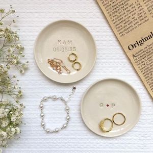 May include: Two cream-coloured ceramic ring dishes with jewellery. One dish has the initials "K&M" and the date "06.08.25". The other dish has the initials "G + P". A pearl bracelet and gold earrings are also visible.