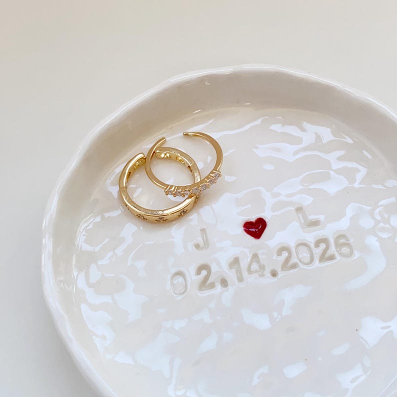 May include: Two gold-colored rings with decorative details rest on a white ceramic dish. One ring has small star-like designs, and the other has a row of small stones. The dish has a red heart and the date 02.14.2026.