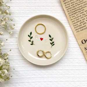 May include: A cream-colored ceramic ring dish with a gold ring and gold hoop earrings. The dish has green leaf designs, the letters "R" and "S", and a red heart. The dish is on a white surface.