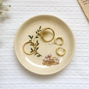 May include: A beige ceramic trinket dish with gold jewelry. The dish features a green leaf design and the initials "G + F". Gold rings, hoop earrings, and a pearl necklace are arranged on the dish.