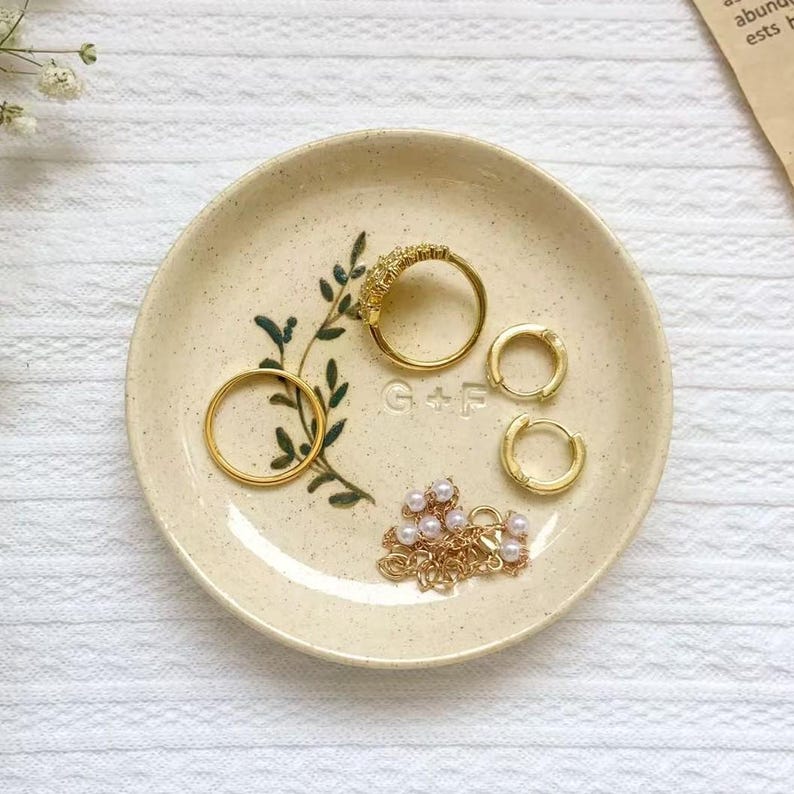 May include: A beige ceramic trinket dish with gold jewelry. The dish features a green leaf design and the initials "G + F". Gold rings, hoop earrings, and a pearl necklace are arranged on the dish.