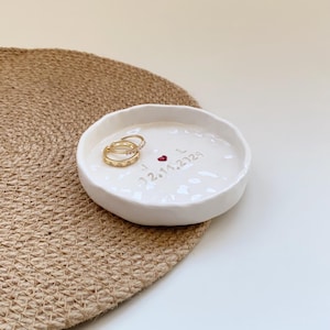 May include: A white ceramic ring dish with a wavy edge, holding two gold rings. The dish is inscribed with the date "02.14.2023", a red heart, and the initials "J L". The dish sits on a woven mat.