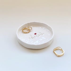 Può includere: Un portanelli in ceramica bianca con superficie strutturata. Due anelli d'oro con piccoli abbellimenti poggiano all'interno del piatto, insieme a un terzo anello d'oro. Il piatto è inciso con le iniziali "J L", un cuore rosso e la data "02.14.2023".