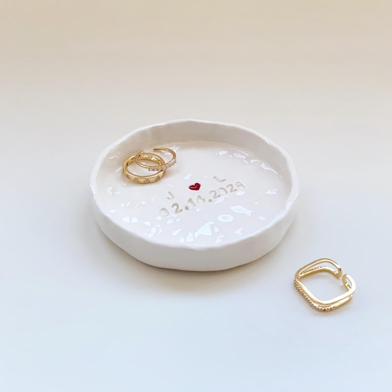 May include: A white ceramic ring dish with a textured surface. Two gold rings with small embellishments rest inside the dish, along with a third gold ring. The dish is inscribed with the initials "J L", a red heart, and the date "02.14.2023".