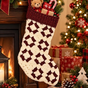 May include: A crochet Christmas stocking featuring a cream and dark red granny square pattern with a solid dark red cuff. The stocking is filled with a small teddy bear, a wrapped gift box, and a candy cane, hanging near a fireplace and tree.