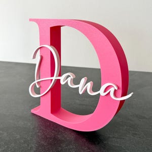 Puede incluir: Una letra "D" rosa brillante, independiente, con el nombre "Dana" en escritura blanca. El diseño en capas crea un efecto 3D, perfecto para decoración personalizada o regalos. La letra es un acento audaz y llamativo.