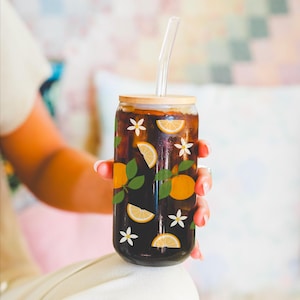 May include: A glass reusable can with a wooden lid and a straw. The can is filled with a dark liquid and has a pattern of orange slices, green leaves, and white flowers.