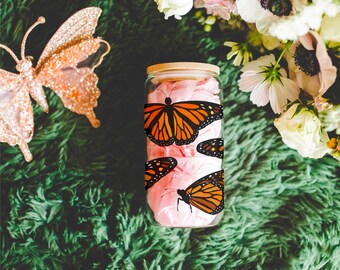 May include: A clear glass jar with a bamboo lid, wrapped with a band of orange and black monarch butterfly illustrations. The jar is filled with pink tissue paper. A decorative rose gold butterfly and flowers are in the background.