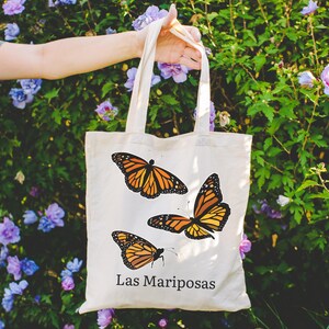 May include: Cream-colored tote bag featuring three orange and black monarch butterflies. The words "Las Mariposas" are printed in black below the butterflies. The bag is held in front of a background of green foliage and purple flowers.