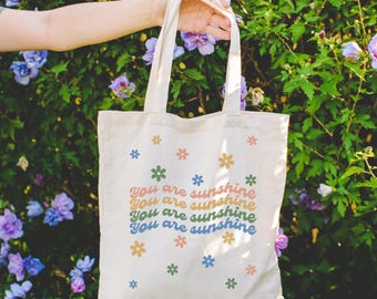 May include: Cream-colored tote bag with the text "You are sunshine" in colorful lettering and small flower graphics. The bag is held up against a backdrop of green foliage and purple flowers.