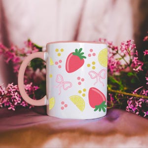 May include: A white ceramic mug with a pink rim and handle. The mug features a repeating pattern of pink bows, yellow lemons, and red strawberries on a white background.