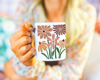 May include: White ceramic mug with a floral pattern in brown, orange, and pink, with green stems and leaves. The mug is held by a person wearing a floral patterned shirt.