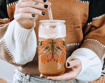 May include: A clear glass tumbler with a bamboo lid and a glass straw. The tumbler features a butterfly design made of orange, green, and brown flowers. The glass is filled with a beverage.