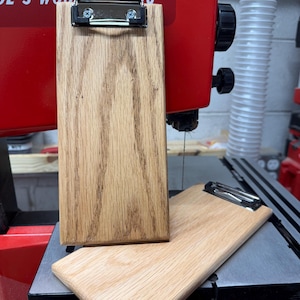 May include: Two wooden clipboards with silver clips. One clipboard is upright, the other is lying down. The clipboards are made of light-colored wood with a visible grain. The background includes a red woodworking machine.