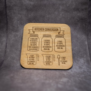 May include: Wooden kitchen conversion chart with engraved measurements. Includes conversions for gallons, quarts, pints, cups, ounces, and liters. Features illustrations of jars, measuring cups, and a spoon. The text "KITCHEN CONVERSION" is at the top.