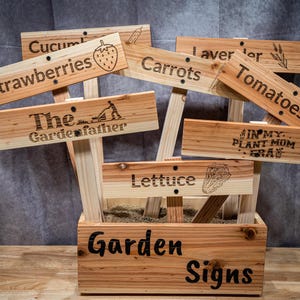 Könnte beinhalten: Gartenschilder aus Holz mit graviertem Text und Illustrationen. Die Schilder sind für verschiedene Pflanzen, darunter Erdbeeren, Karotten und Tomaten. Die Schilder werden in einer Holzkiste mit der Aufschrift "Garden Signs" angezeigt.