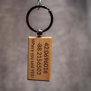 May include: A wooden rectangular keychain with engraved text and numbers. The text reads "Where you said YES!" and includes the coordinates 42.0696016, -88.2156503. The keychain is attached to a metal keyring and chain.