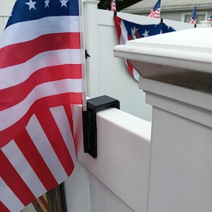 May include: A black flag holder is attached to a white fence, displaying an American flag with red and white stripes and blue stars. Other American flags are visible in the background, creating a patriotic theme.