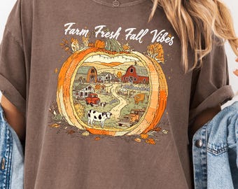 May include: Espresso-colored t-shirt with a fall-themed graphic. The graphic features a pumpkin with a farm scene inside, including a barn, a cow, and the text "Farm Fresh Fall Vibes". The shirt is paired with a light blue denim jacket.