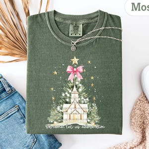 May include: A moss green t-shirt featuring a Christmas tree design with a pink bow, gold stars, and a church. The text "Oh come, let us adore Him" is printed at the bottom. A silver necklace is draped across the shirt.