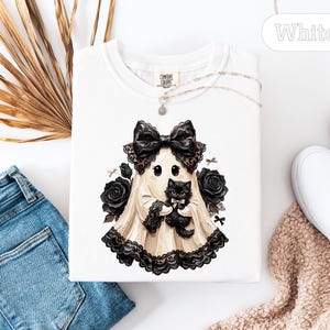 May include: A white t-shirt featuring a graphic of a ghost wearing a large black lace bow and holding a fluffy black kitten. The ghost is surrounded by black roses and small bows. The shirt has a crew neckline and a Comfort Colors brand label.