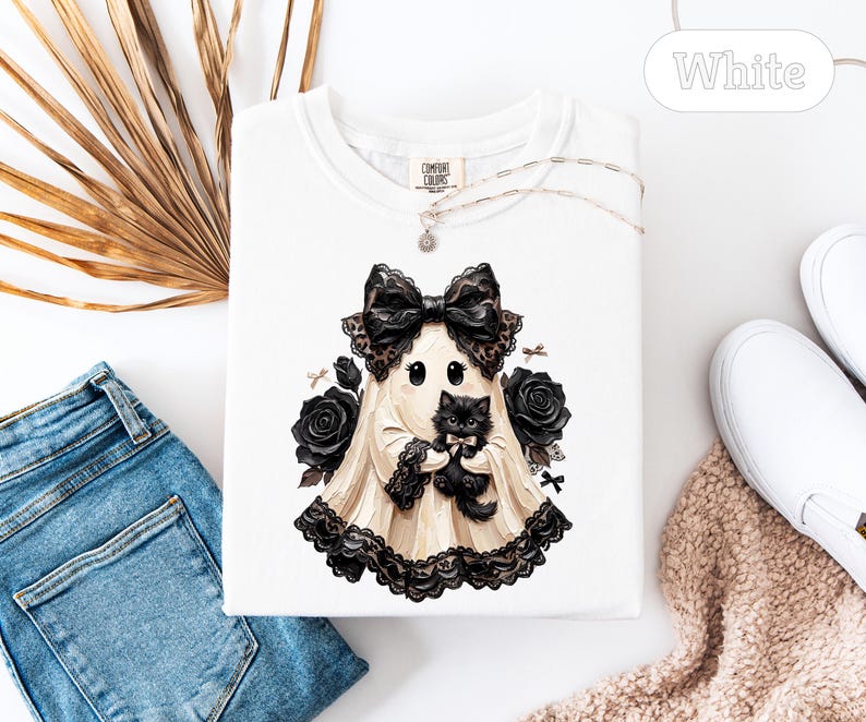 May include: A white t-shirt featuring a graphic of a ghost wearing a large black lace bow and holding a fluffy black kitten. The ghost is surrounded by black roses and small bows. The shirt has a crew neckline and a Comfort Colors brand label.