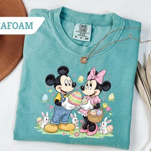 May include: Seafoam green t-shirt featuring Mickey and Minnie Mouse holding Easter eggs. The graphic includes Easter eggs, bunnies, and flowers. A gold necklace is draped across the shirt. The shirt is labeled "Seafoam".