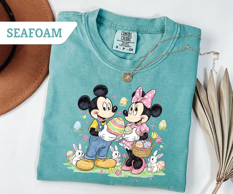 May include: Seafoam green t-shirt featuring Mickey and Minnie Mouse holding Easter eggs. The graphic includes Easter eggs, bunnies, and flowers. A gold necklace is draped across the shirt. The shirt is labeled "Seafoam".