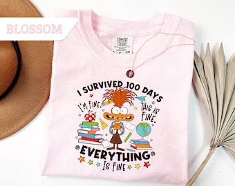 May include: A light pink t-shirt with the text "I SURVIVED 100 DAYS, I'M FINE, THIS IS FINE, EVERYTHING IS FINE" and a cartoon character illustration. The shirt has a crew neck and short sleeves. A gold necklace is draped across the shirt.