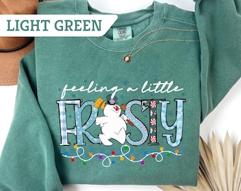 Puede incluir: Sudadera verde claro con la frase "feeling a little FROSTY" en letras blancas y azules. El diseño incluye un muñeco de nieve, copos de nieve y una guirnalda de luces de colores. Sudadera de la marca Comfort Colors.