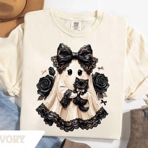 May include: An ivory t-shirt featuring a graphic of a ghost wearing a large black lace bow and holding a fluffy black kitten. The ghost is surrounded by black roses and small black bows, with a black lace trim along the bottom hem of the ghost.
