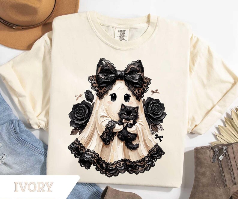 May include: An ivory t-shirt featuring a graphic of a ghost wearing a large black lace bow and holding a fluffy black kitten. The ghost is surrounded by black roses and small black bows, with a black lace trim along the bottom hem of the ghost.