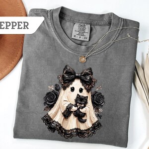 May include: A pepper-colored t-shirt featuring a graphic of a ghost wearing a large black bow and lace trim, holding a small black kitten. The ghost is surrounded by black roses. The shirt has a crew neckline and a Comfort Colors brand label.