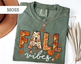 May include: A moss green t-shirt with the words "FALL vibes" in a fall-themed design. The word "FALL" is in orange and brown with fall leaves and pumpkin details. A silver necklace is draped across the shirt.