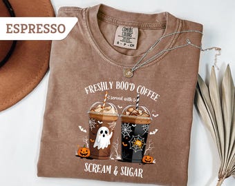 May include: Espresso-colored t-shirt featuring a Halloween-themed graphic. The design includes two iced coffee cups with spooky decorations, a ghost, spiderwebs, and pumpkins. The text reads "Freshly Boo'd Coffee served with Scream & Sugar."