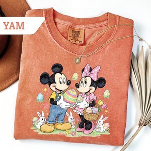 May include: Coral-colored t-shirt featuring Mickey and Minnie Mouse holding a large Easter egg. The graphic includes Easter eggs, bunnies, and flowers. The shirt has a crew neck and short sleeves. A gold necklace is draped over the shirt.