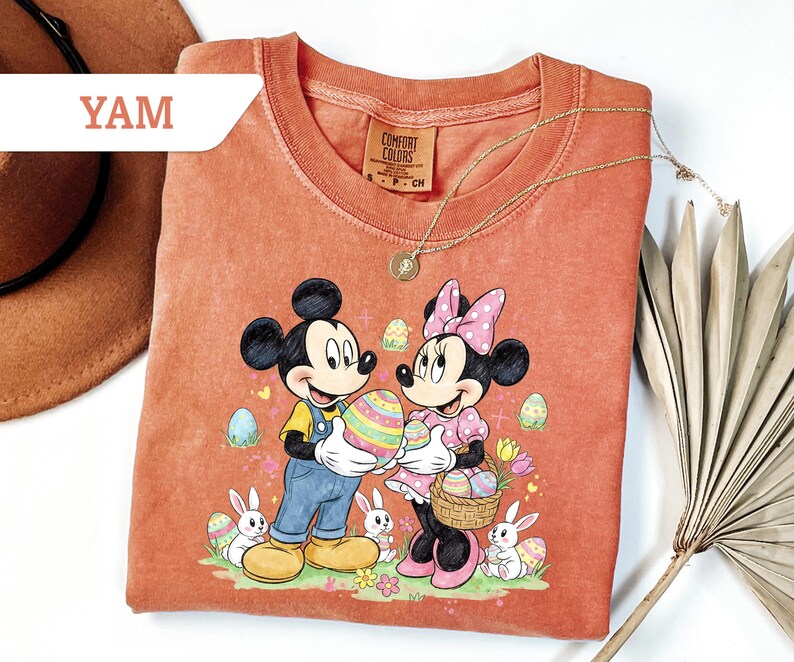 May include: Coral-colored t-shirt featuring Mickey and Minnie Mouse holding a large Easter egg. The graphic includes Easter eggs, bunnies, and flowers. The shirt has a crew neck and short sleeves. A gold necklace is draped over the shirt.
