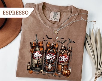 May include: An espresso-colored t-shirt featuring a graphic of three Dr. Pepper cans decorated with Halloween themes, including a haunted house, a ghost, and a pumpkin. The cans are topped with plaid bows and straws, surrounded by bats and leaves.
