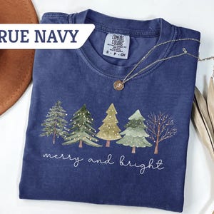 May include: A navy blue t-shirt with a Christmas tree design and the words "merry and bright" in white script. The shirt has a crew neck and short sleeves. A gold necklace is draped across the shirt. The words "True Navy" are in a white banner.