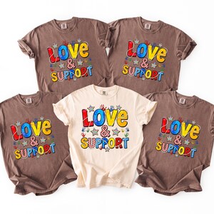 May include: Five t-shirts with the words "Love & Support" in colorful letters and designs. The shirts are in various colors, including brown and cream. The design includes stars and handprints.