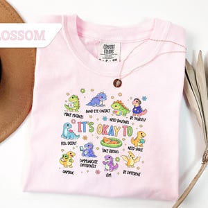 May include: A light pink Comfort Colors t-shirt featuring a colorful graphic of cartoon dinosaurs with the text "It's Okay To" and various phrases. The shirt has a crew neck and short sleeves. A gold necklace is draped over the shirt.