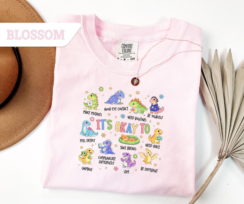May include: A light pink Comfort Colors t-shirt featuring a colorful graphic of cartoon dinosaurs with the text "It's Okay To" and various phrases. The shirt has a crew neck and short sleeves. A gold necklace is draped over the shirt.