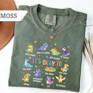 May include: Moss green t-shirt with a crew neck and short sleeves. The shirt features a colorful graphic of cartoon dinosaurs with the text "It's Okay To" and various phrases. A gold necklace with a pendant is draped across the shirt.