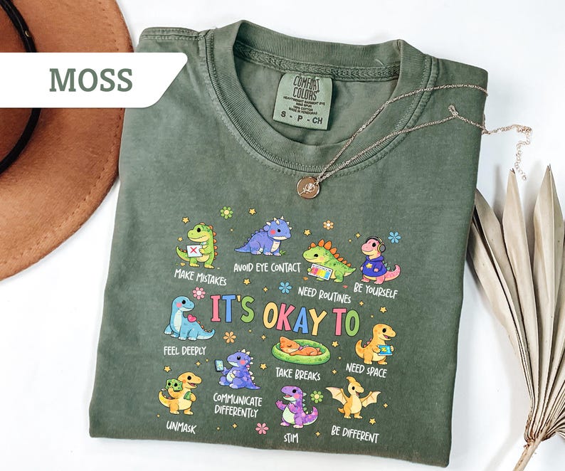 May include: Moss green t-shirt with a crew neck and short sleeves. The shirt features a colorful graphic of cartoon dinosaurs with the text "It's Okay To" and various phrases. A gold necklace with a pendant is draped across the shirt.