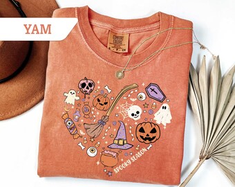 May include: Orange-red Halloween-themed t-shirt with a heart-shaped design. The design includes a skull, pumpkin, witch's hat, broom, and the words "Spooky Season". A gold necklace is draped over the shirt.