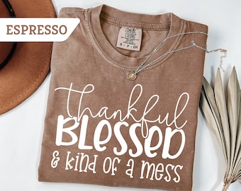 May include: Espresso-colored t-shirt with the words "Thankful, Blessed & kind of a mess" in white script. The shirt has a crew neck and short sleeves. A silver necklace with a pendant is draped on the shirt.