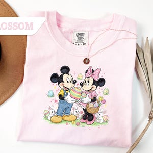 May include: A light pink t-shirt featuring Mickey and Minnie Mouse holding a decorated Easter egg and basket. The graphic includes Easter eggs, bunnies, and flowers. The word "BLOSSOM" is in a white rectangle in the upper left corner.