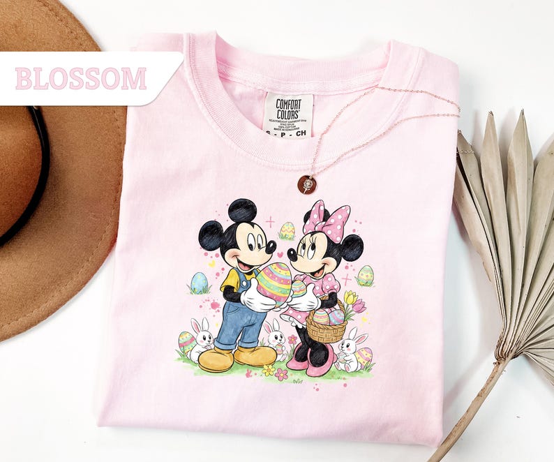 May include: A light pink t-shirt featuring Mickey and Minnie Mouse holding a decorated Easter egg and basket. The graphic includes Easter eggs, bunnies, and flowers. The word "BLOSSOM" is in a white rectangle in the upper left corner.