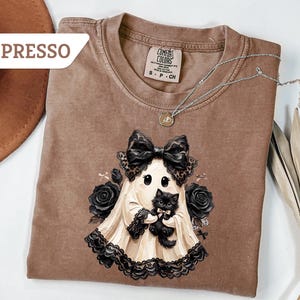 May include: An espresso-colored t-shirt featuring a graphic of a ghost wearing a large black bow and lace trim, holding a small black kitten. The ghost is surrounded by black roses. The shirt has a Comfort Colors label at the inner neckline.