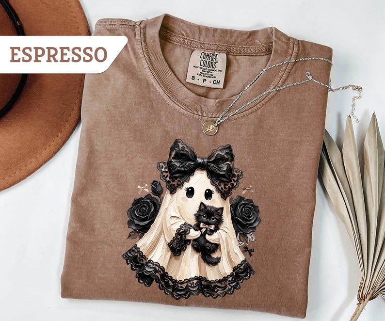 May include: An espresso-colored t-shirt featuring a graphic of a ghost wearing a large black bow and lace trim, holding a small black kitten. The ghost is surrounded by black roses. The shirt has a Comfort Colors label at the inner neckline.